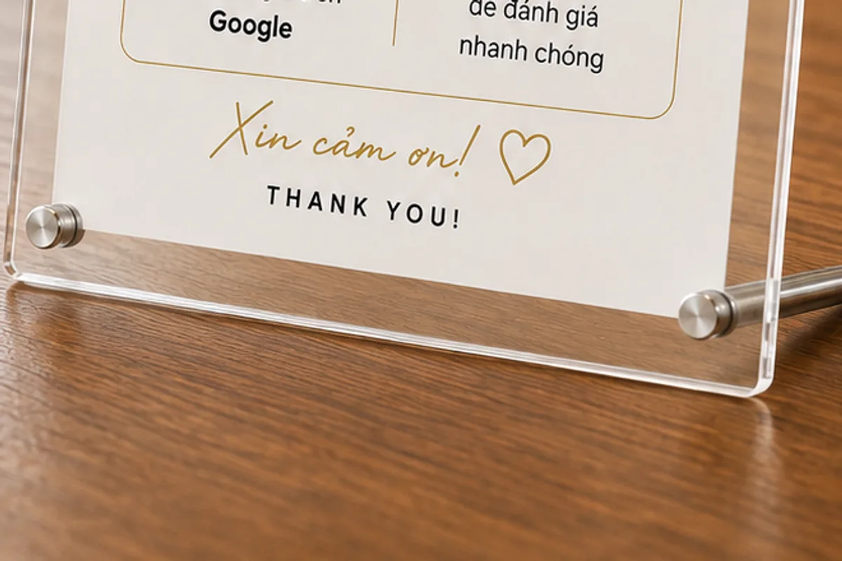 Cận cảnh chân standee: hai tấm acrylic trong kẹp bản in ở giữa, bắt bằng ốc trụ inox tròn đầu, phía dưới là một thanh kim loại tròn đỡ ngang, đặt trên mặt bàn gỗ sẫm