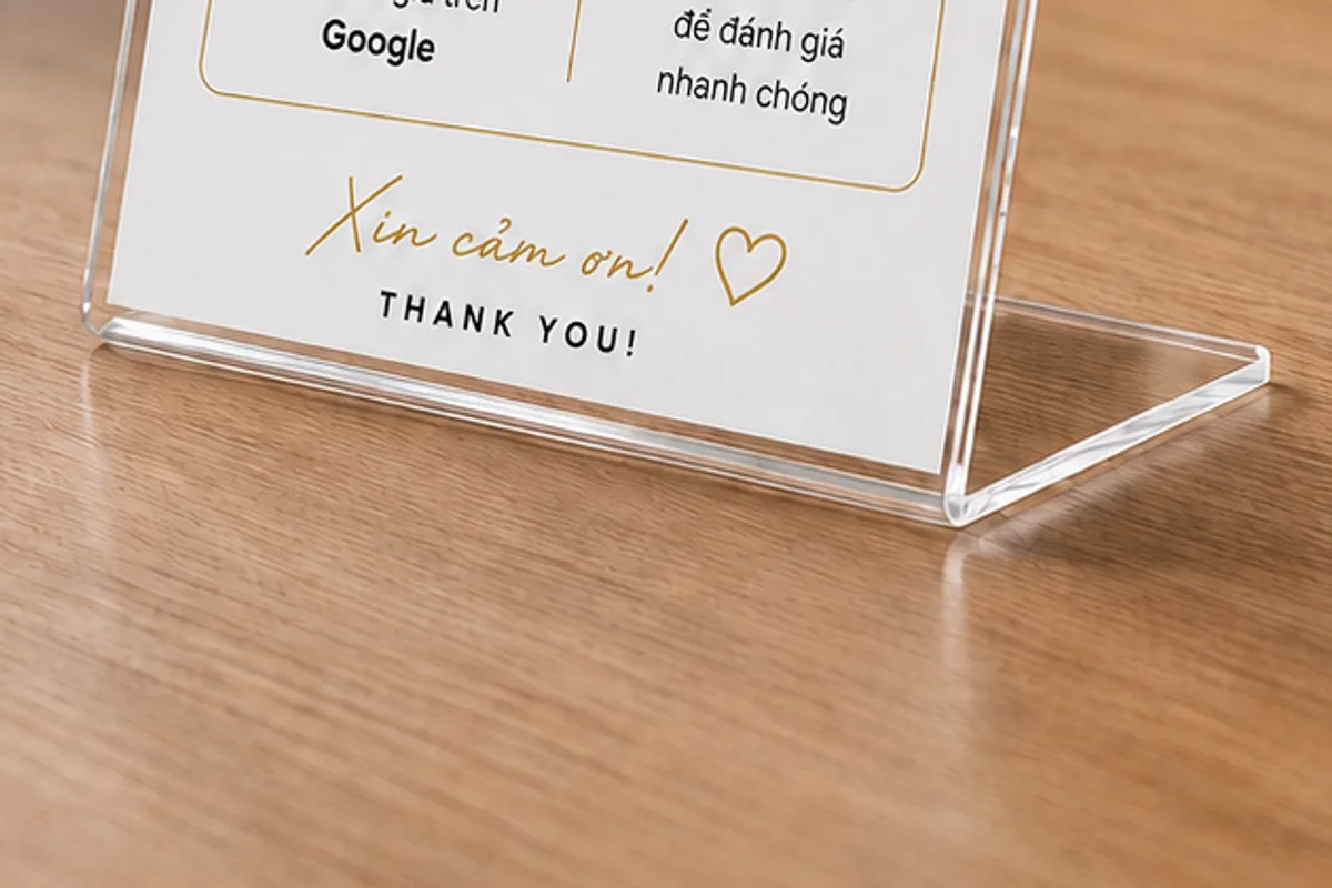 Cận cảnh chân standee: tấm acrylic trong suốt uốn gập thành chữ L liền một khối, mép gập bo tròn, đặt trên mặt bàn gỗ sáng